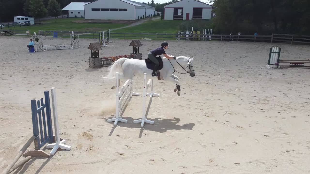 Una toma de dron volando junto a un caballo blanco y un jinete mientras saltan múltiples obstáculos en una pista al aire libre.