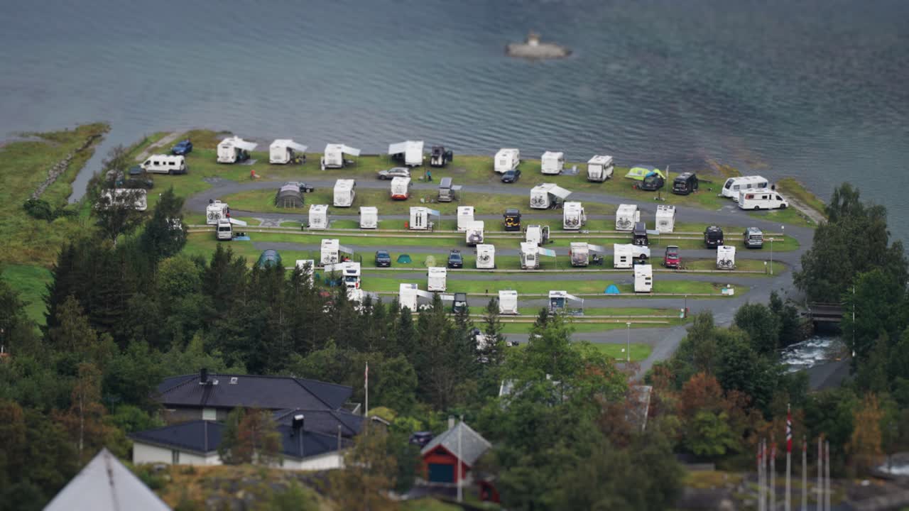 A campsite by the water on the shores of the Geiranger fjord, featuring rows of RVs and tents surrounded by lush greenery. A timelapse video.
