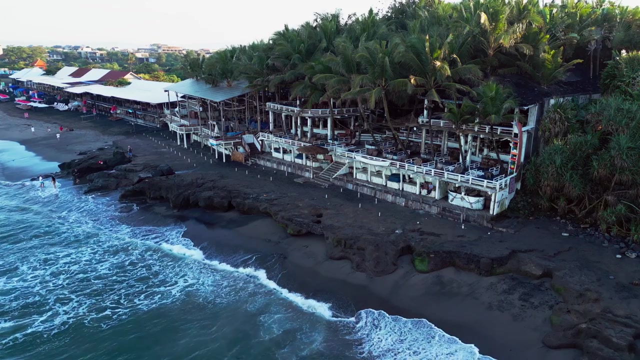 Veduta aerea con drone del beach club La Brisa, una struttura all'aperto in legno di recupero con ringhiere in legno bianco sotto alte palme da cocco su una costa di roccia vulcanica scura con onde dell'oceano che si infrangono a Canggu, isola di Bali