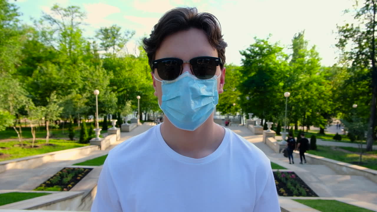 Giovane uomo con camicia bianca e occhiali da sole che si toglie una mascherina medica protettiva dal viso mentre guarda la fotocamera. Giornata di sole al parco. Idea sul Coronavirus