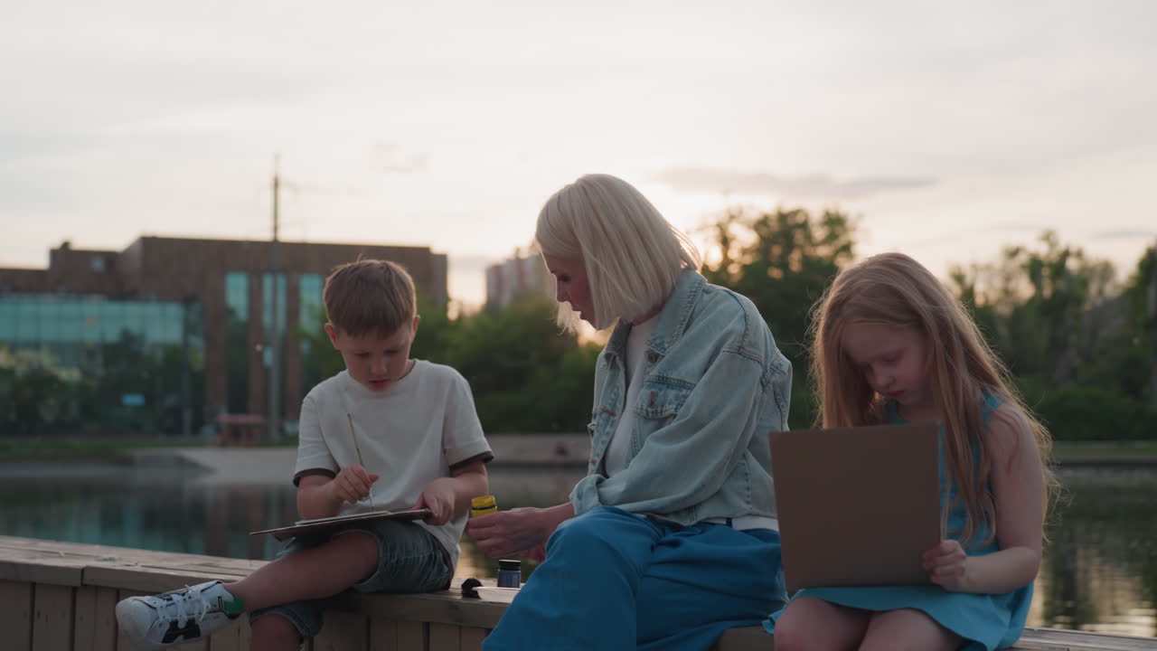 young mom seated on wooden dock between two kids guiding painting lesson with paint and brush on paper by shimmering pool under sunset glow, siblings focused as mother demonstrates strokes