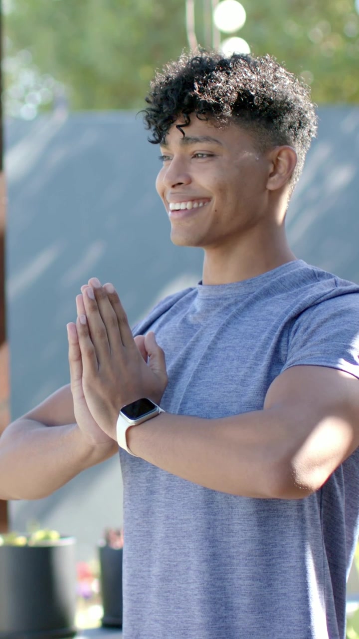 Video vertical: Hombre sonriente con camiseta gris haciendo namaste, girando a la derecha al iniciar la cámara, en un jardín.