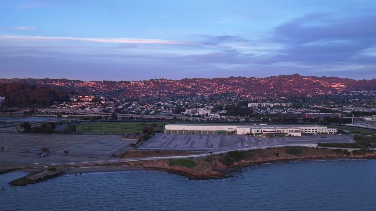Vista aerea che sorvola le acque calme della baia di San Francisco, mostrando la penisola di Albany Bulb e le lontane colline di East Bay che brillano con l'ultima luce di un bellissimo tramonto californiano.