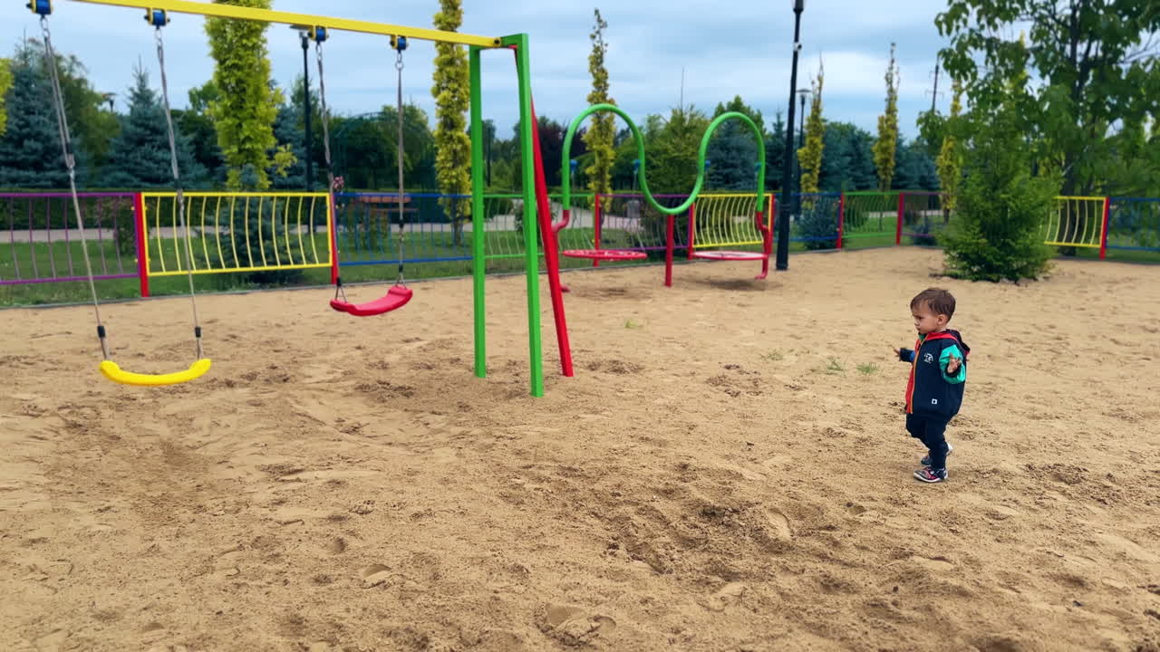 Adorable child walks by the sand on the playground holding hands to the sides. Kid comes up to the swing.