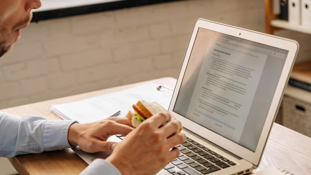 Uomo alla scrivania che addenta un panino mentre digita al portatile, modifica un documento e poi invia un modulo per lavoro.