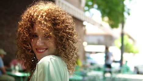 hermosa chica de cabello rojo con el peinado perfecto - rizos al aire libre, sonriendo y posando ante la cámara, dando la vuelta con una terraza de café al aire libre en el fondo. cámara lenta