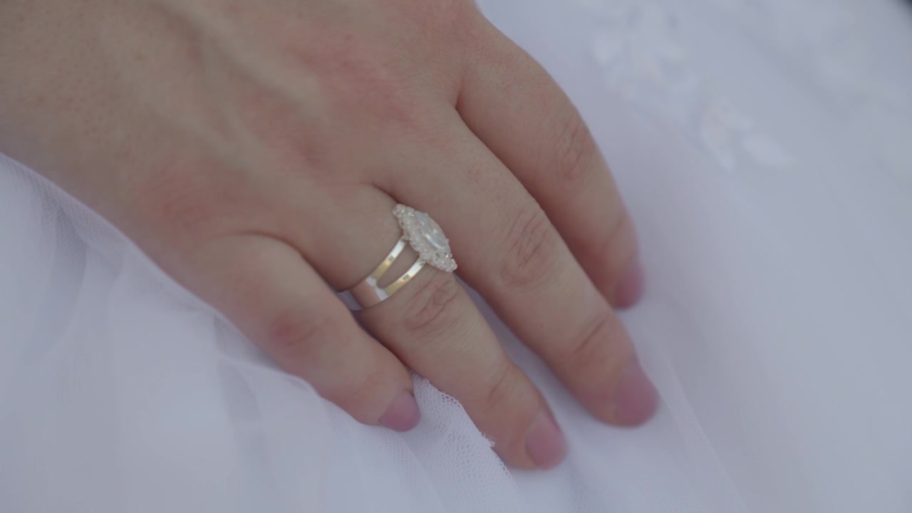 Mano della sposa che poggia su un abito da sposa bianco, mostrando un anello di fidanzamento e una fede nuziale scintillanti mentre la mano si distende.