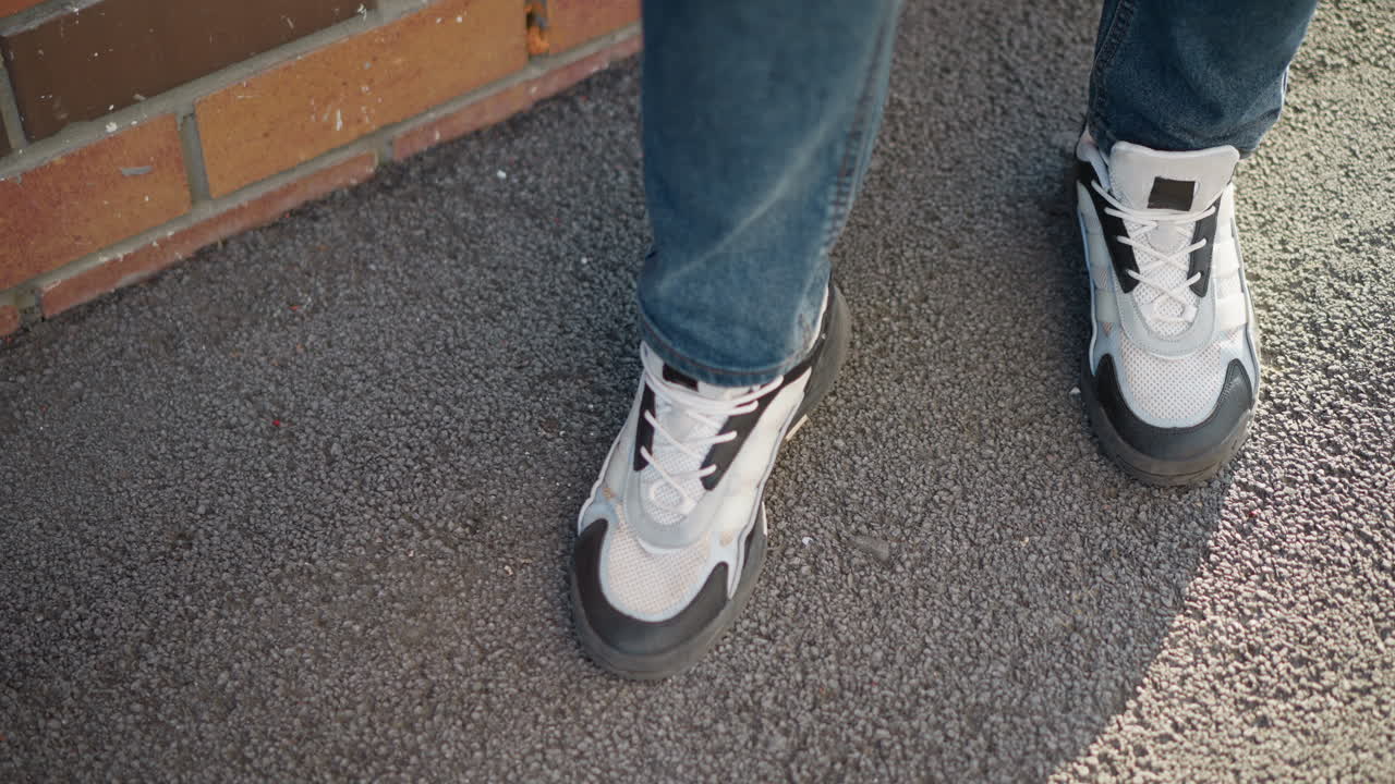 top down view of man in jeans and sneakers stepping on nearly finished cigarette butt on asphalt pavement next to brick wall in daylight, suggesting end of smoke break in urban outdoor setting