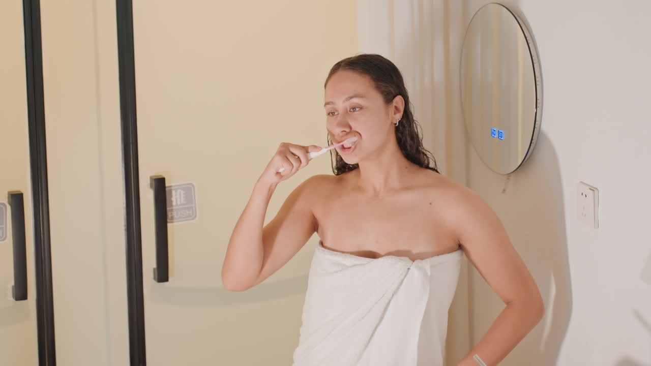 Confident hotel guest prepping for day while brushing teeth in towel, business traveler styling and preparing outfit, mirror reflection shows composed posture and modern bathroom amenities, smooth