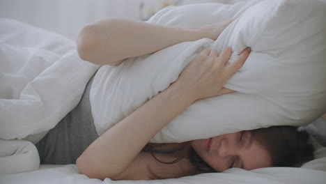 young woman laying in bed suffers from noise generated by the construction site outside the window. woman covering her ears with pillow.