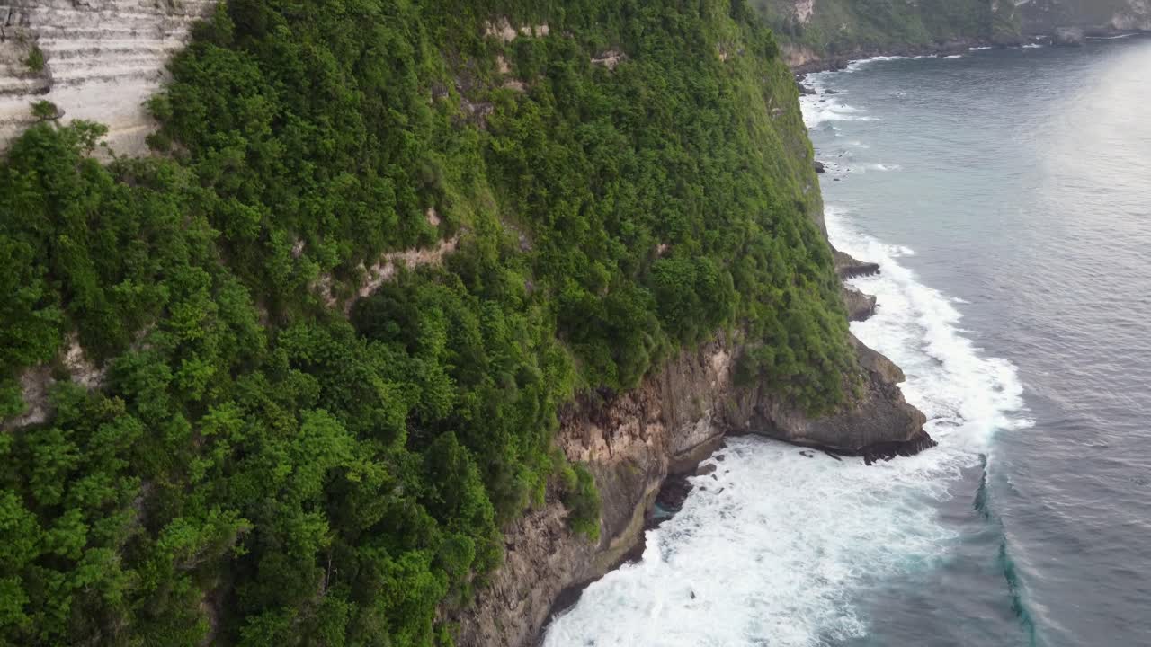 Aerial Ascending Reveal Tropical seaside of Nusa Penida Island high steep coastal cape edge overlooking the waves of indian ocean crashing down the green vegetated cliff, Indonesia