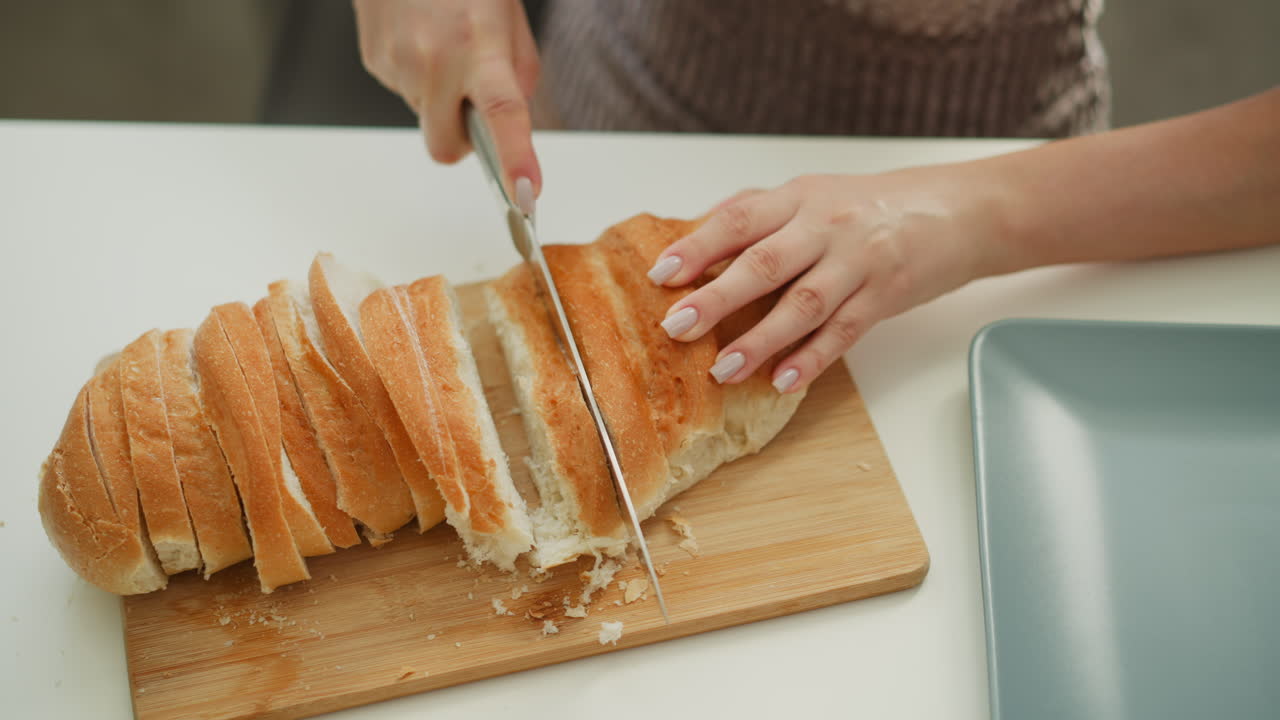 High angle hand view of saucier slicing loaf of bread on wooden board with knife on table next to egg banana and bowls in modern kitchen with sunny morning light streaming through window behind