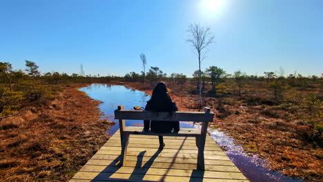 solo woman relaxes sitting on wooden bench standing up to look out over wetland plains at midday