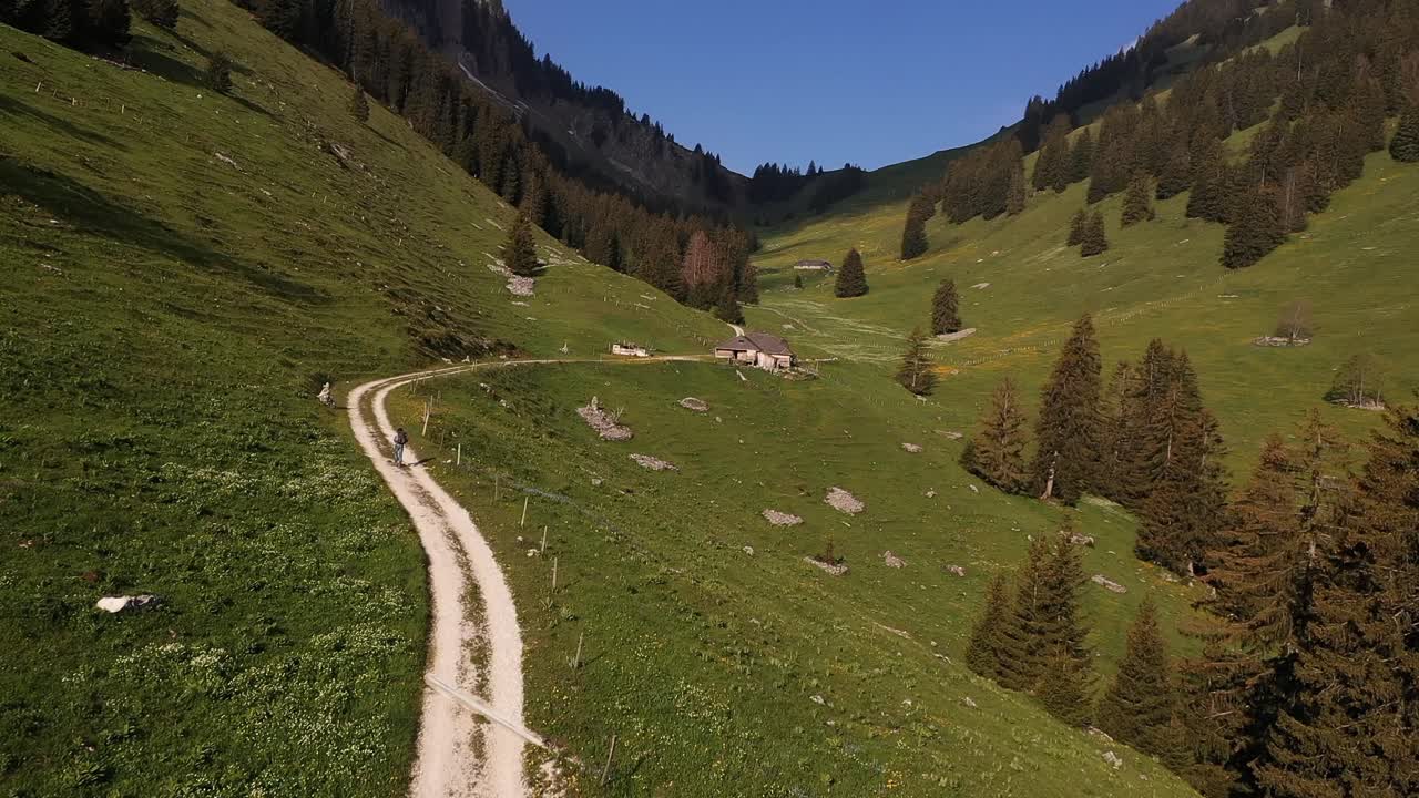 Aerial, drone shot backpacking trekking alone on mountain valley trail towards a house