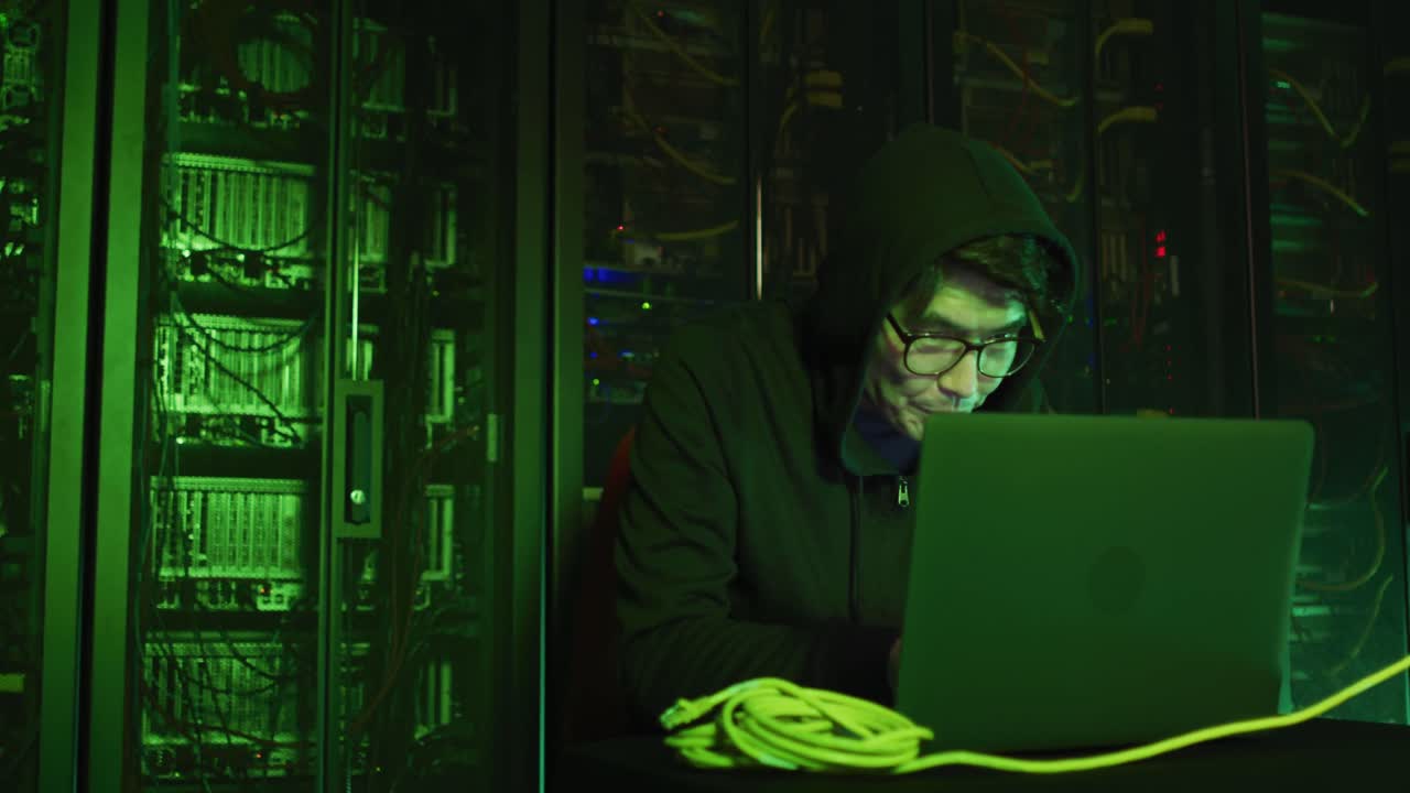 Asian male hacker in hoodie using laptop by computer servers