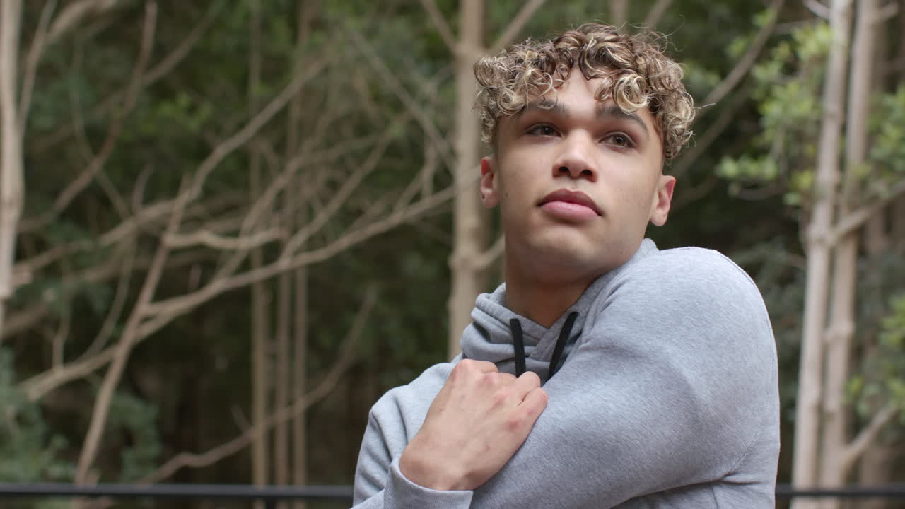 Young man in hoodie stretching outdoors, looking thoughtful in forest setting, copy space
