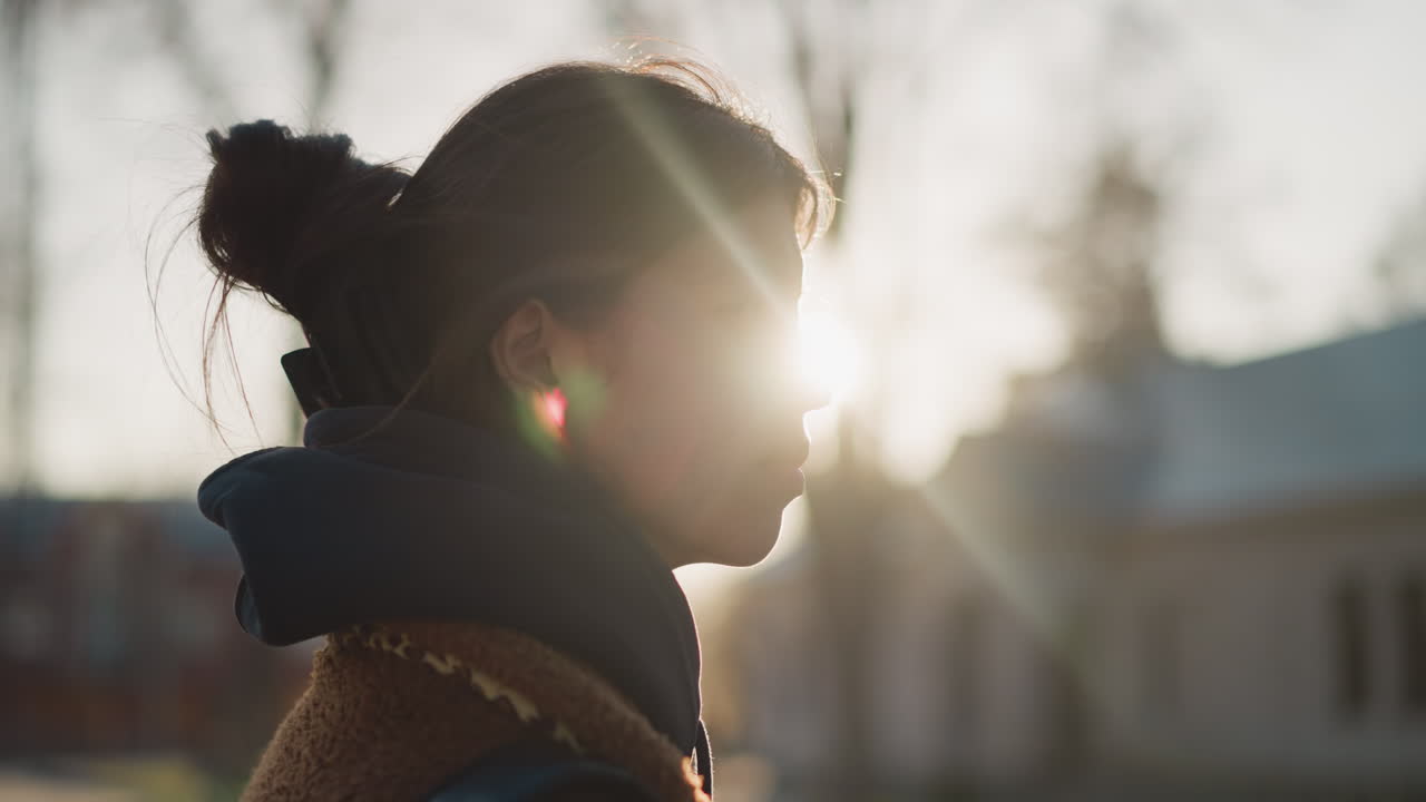 A close-up of a girl walking with a faint and sad expression, captured against the backdrop of a setting sun. The soft light of the sunset adds a reflective and emotional atmosphere