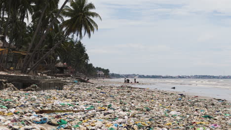 tropical beach polluted with plastic trash, locals cleaning up in the distance