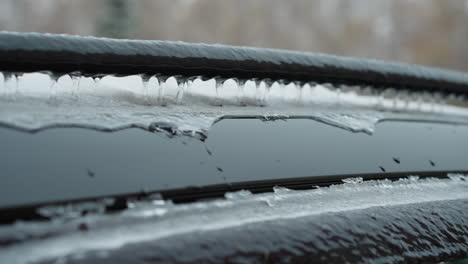 close-up of vehicle roof covered in thick ice and snow, highlighting icicles forming along edges of black metal rail, set against a blurred urban winter background