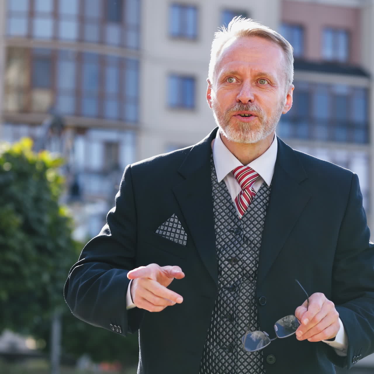 Portrait senior businessman in eyeglasses on the building background in the city. Serious bearded man with grey hair in suit telling about his business outdoors.