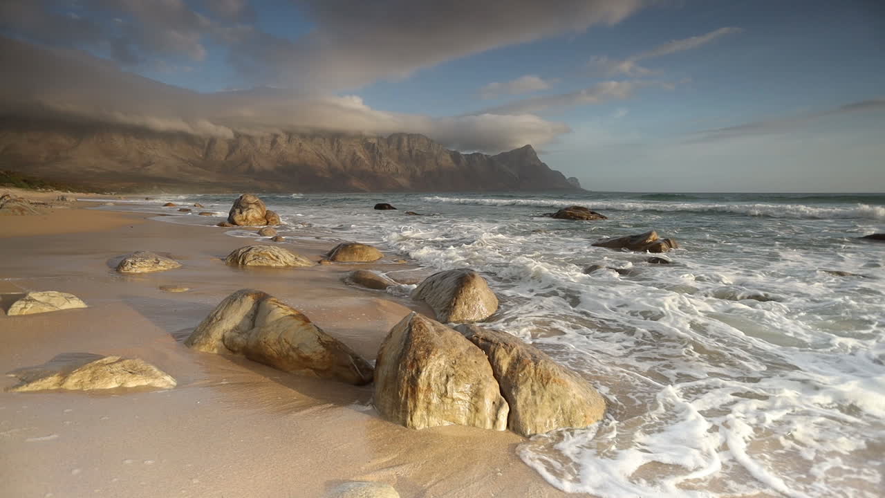 olas rompiendo en una playa rocosa al atardecer con impresionantes montañas en el fondo