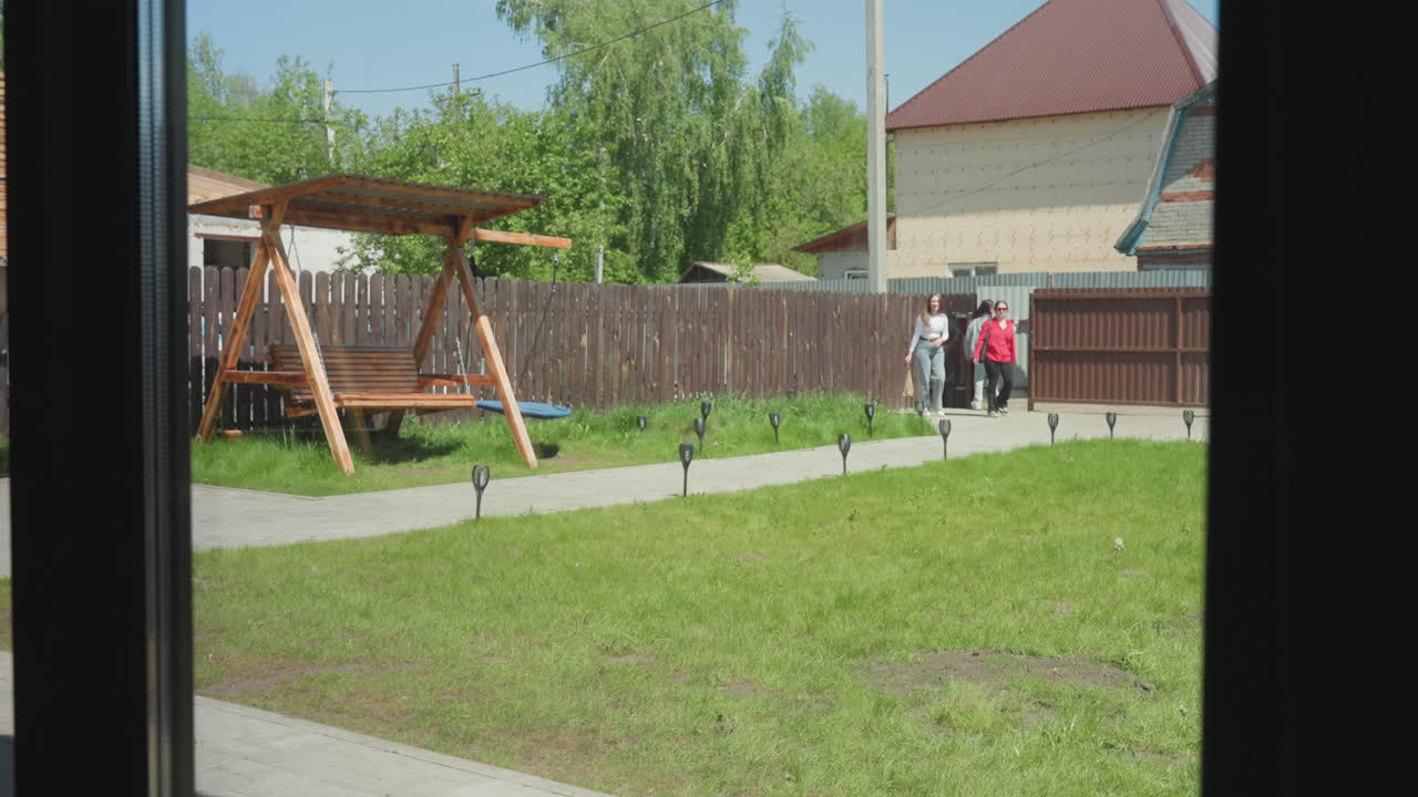 Three ladies seen through glass window entering compound through open gate, walking along paved path with wooden swing, green lawn, and residential houses in background under clear sunny sky