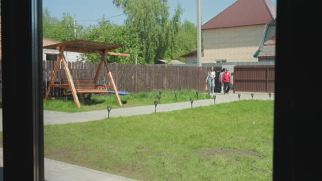 three ladies seen through glass window entering compound through open gate, walking along paved path with wooden swing, green lawn, and residential houses in background under clear sunny sky