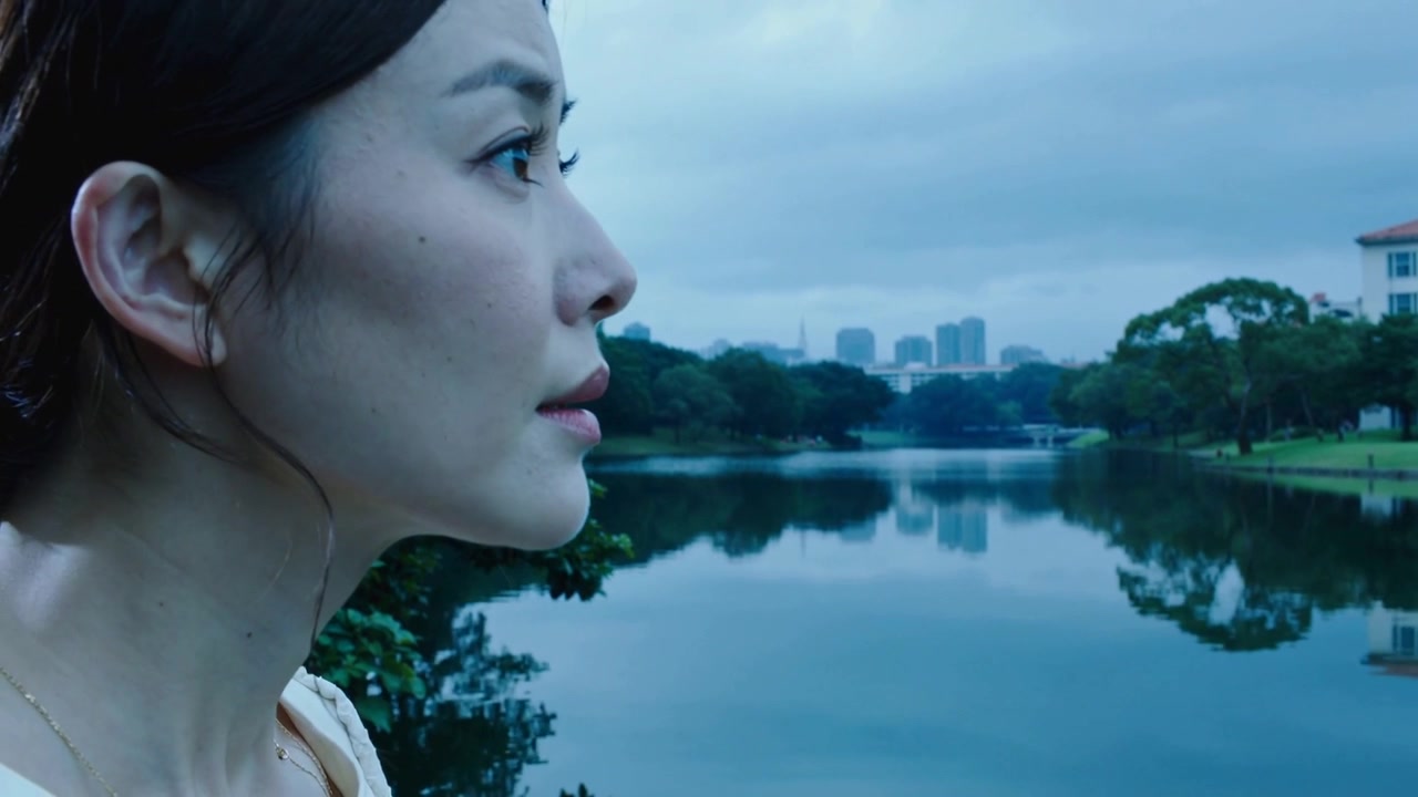 凝视的亚洲女子抬起下巴，转向公园里平静的湖面，沉思着，头上戴着白色发夹。