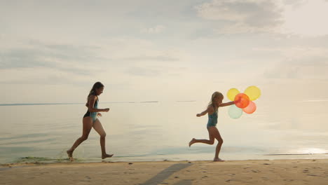 due sorelle che giocano insieme sulla spiaggia una ragazza corre dietro a un'altra tenendo dei palloncini rallentatore s