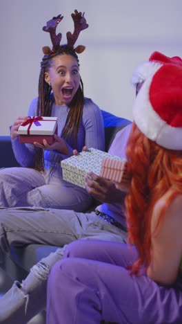 Vertikale-Videostudioaufnahme-Von-Gen-Z-Freunden,-Die-Zu-Weihnachten-Geschenke-überreichen-Und-öffnen,-Sitzend-Auf-Dem-Sofa-Mit-Weihnachtsmütze-Und-Rentiergeweih-2