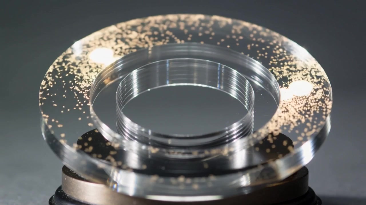 Palco de metal giratório virando um anel de acrílico transparente sob luzes direcionais, com partículas de ouro se movendo.