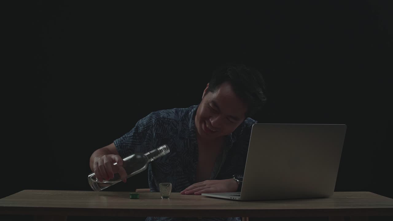 Uomo asiatico che brinda e beve vodka durante una videochiamata al laptop su sfondo nero.