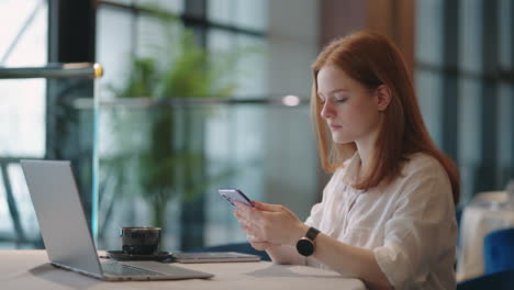 Weibliche-Büroangestellte-Im-Co-Working-Bereich-Sitzen-Am-Tisch-Mit-Laptop-Und-Nutzen-Das-Mobiltelefon-Zum-Versenden-Von-Nachrichten