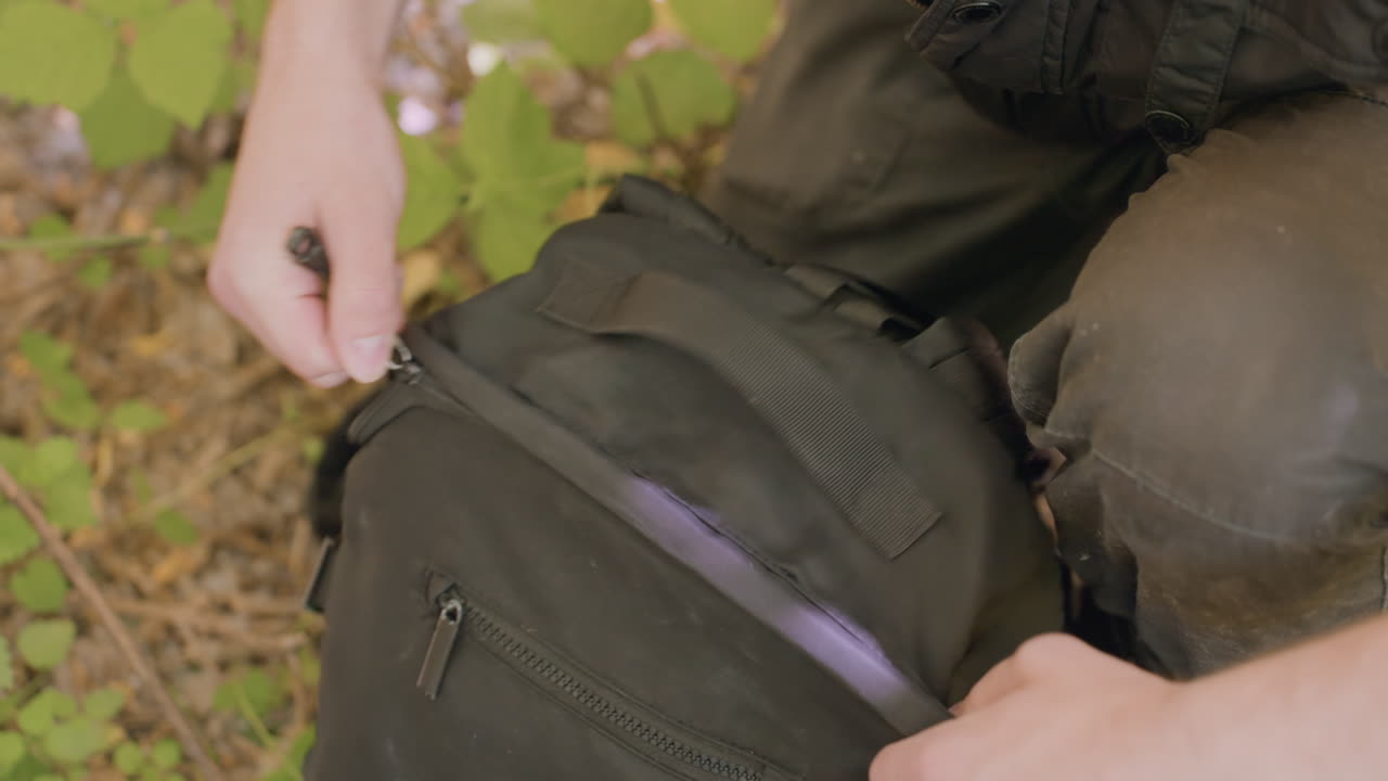 Young man kneels in forest, retrieves item from backbag, places it back, and zips up bag before continuing journey through leafy woodland floor surrounded by twigs, soil, and green plants