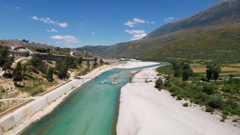 Colorido-Paisaje-Del-Río-Vjosa-Con-Agua-Turquesa-Fluyendo-Sobre-Guijarros,-Montañas-Y-Fondo-De-Cielo-Azul