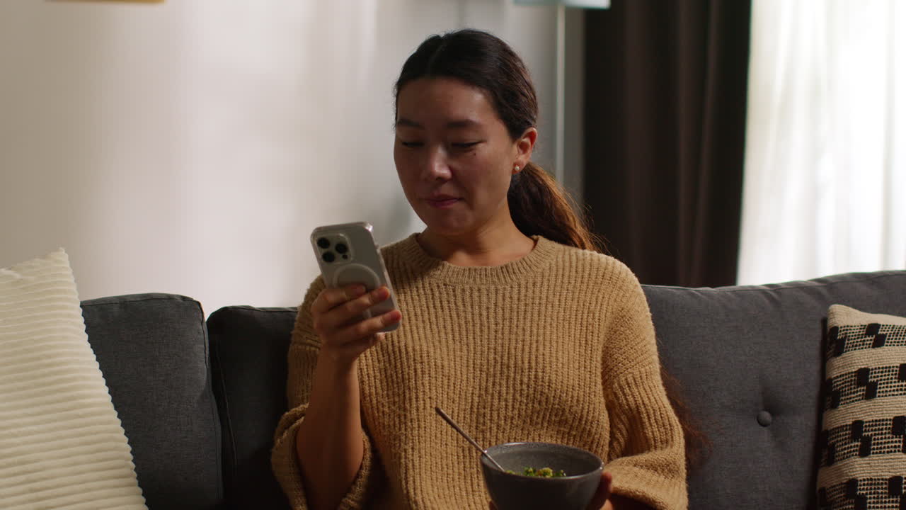 Woman At Home Sitting On Sofa Eating Healthy Vegetarian Or Vegan Meal Of Tabbouleh Salad With Glass Of Wine Looking At Mobile Phone 1