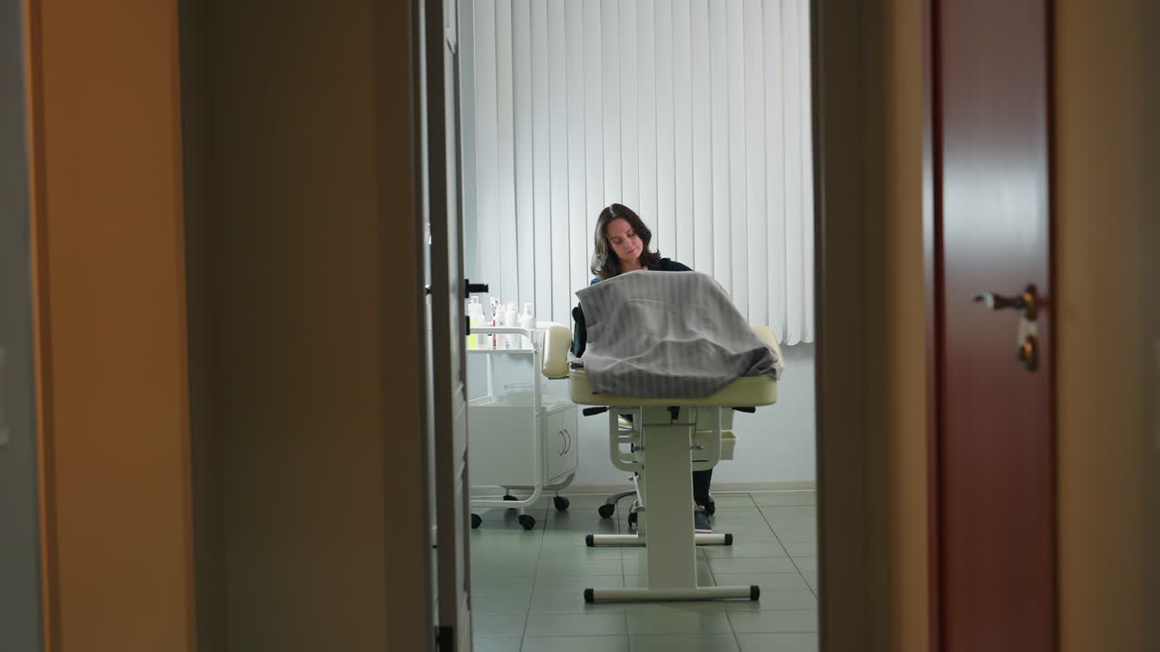 Treatment provider seen through door rendering massage service to customer lying on spa bed in softly lit room with vertical blinds background showcasing expert care and tranquil environment