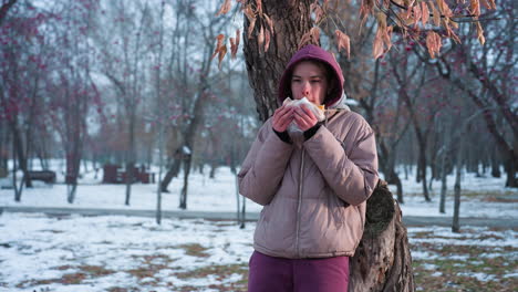 young woman in winter outfit resting on tree eating snack in outdoor park during winter, light reflection from background, enjoying food moment, cozy winter break in nature