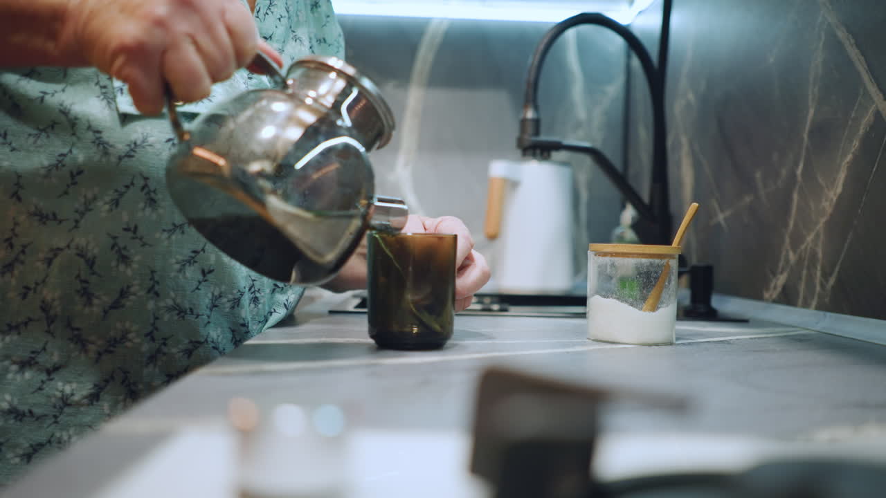 White grandma pouring tea from kettle, steady hands tilting metal pot over green glass mug, steam rising, focus on motion and hospitality, warm domestic scene, mindful morning routine