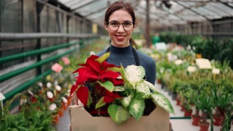 Beautiful-female-gardener-in-black-apron-carrying-cartoon-box-with-flowers-plants