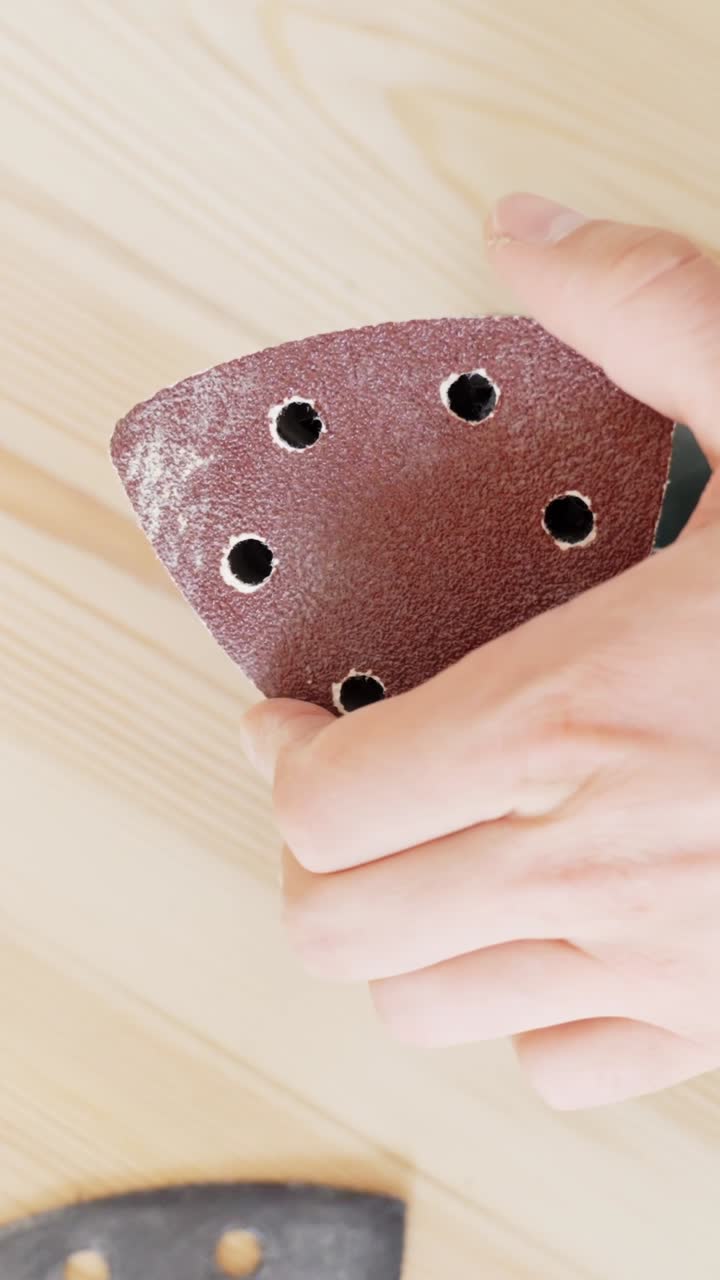 A person’s hands are seen fitting a perforated sanding disc onto a triangular orbital sander, preparing it for wood finishing work on a smooth wooden board in a well-lit interior space