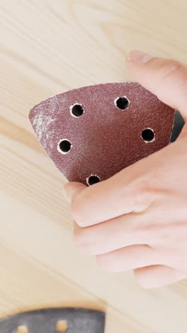a person’s hands are seen fitting a perforated sanding disc onto a triangular orbital sander, preparing it for wood finishing work on a smooth wooden board in a well-lit interior space