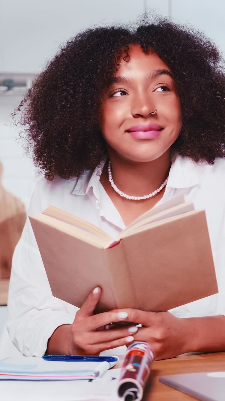 Giovane donna afroamericana positiva e sognante legge un libro seduta al tavolo.