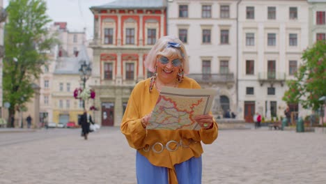 Elderly-stylish-tourist-granny-woman-walking-along-street,-looking-for-way-using-paper-map-in-city