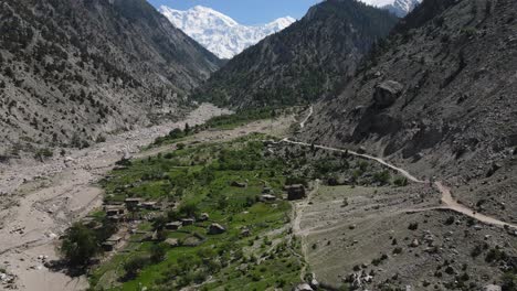 paisaje del norte de pakistán, vista aérea del pueblo y camino de tierra bajo colinas rocosas y picos montañosos nevados
