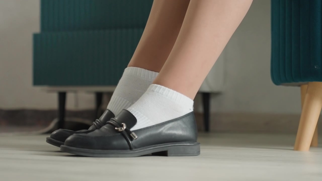 Donna caucasica che si mette le scarpe in casa, primo piano di calzini bianchi che scivolano in mocassini neri su pavimento laminato, mano che stabilizza il piede, soggiorno accogliente, atmosfera domestica calma, interno pulito e ordinato.