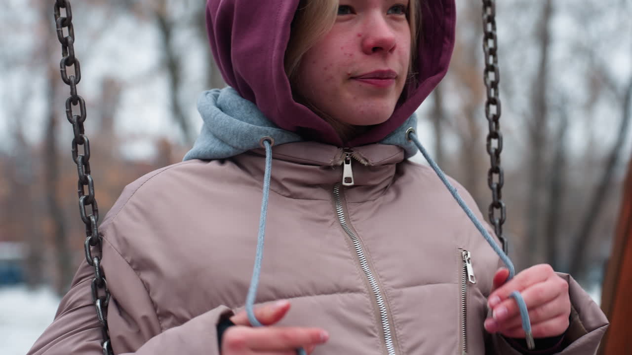 mujer joven en traje de invierno sentada en el columpio, jugando con encaje de capucha en clima frío, suelo cubierto de nieve y árboles desnudos en el fondo