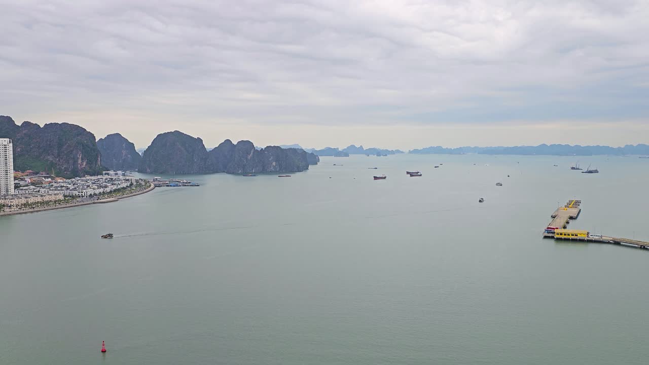 A panoramic view of Ha Long Bay showcases dramatic limestone karsts rising from tranquil waters, with a modern cityscape along the shoreline, scattered boats, distinctive yellow pier in misty bay