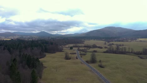 aerial of rural landscape. clouds and mountains in background. flying slowly forwards to forest. empty road leading into woods.
