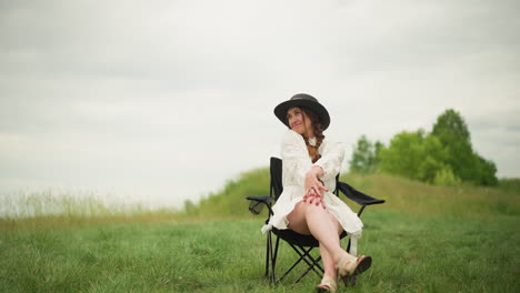 a pretty woman wearing a white dress and a black hat sits on a chair in a grassy field, posing with a relaxed and confident demeanor. a natural and serene backdrop to her stylish appearance